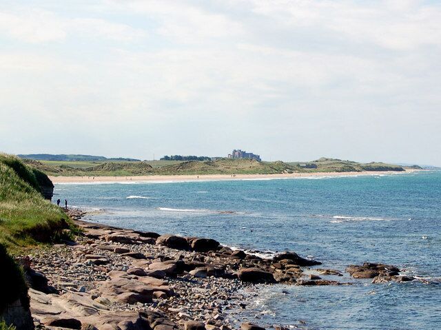 A rocky beach with waves, a distant building, and grassy hills.