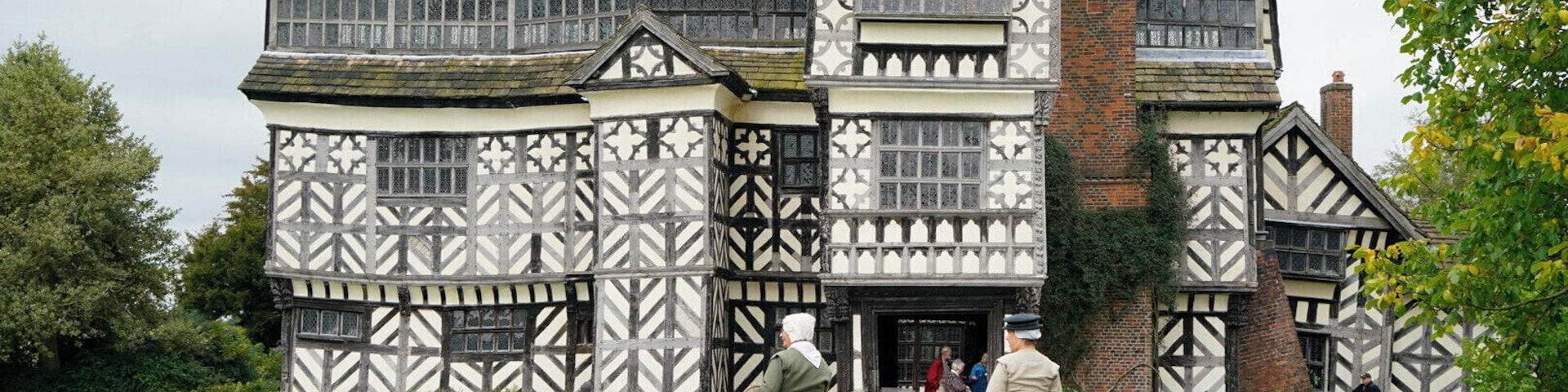 Um edifício em estilo Tudor, com fachada em madeira branca e preta, chaminé de tijolos e um caminho pavimentado que leva à entrada.