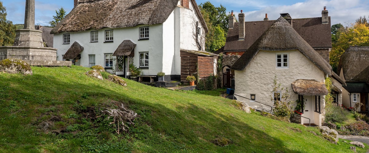 A traditional white house with a thatched roof, a stone monument, and a grassy hill.