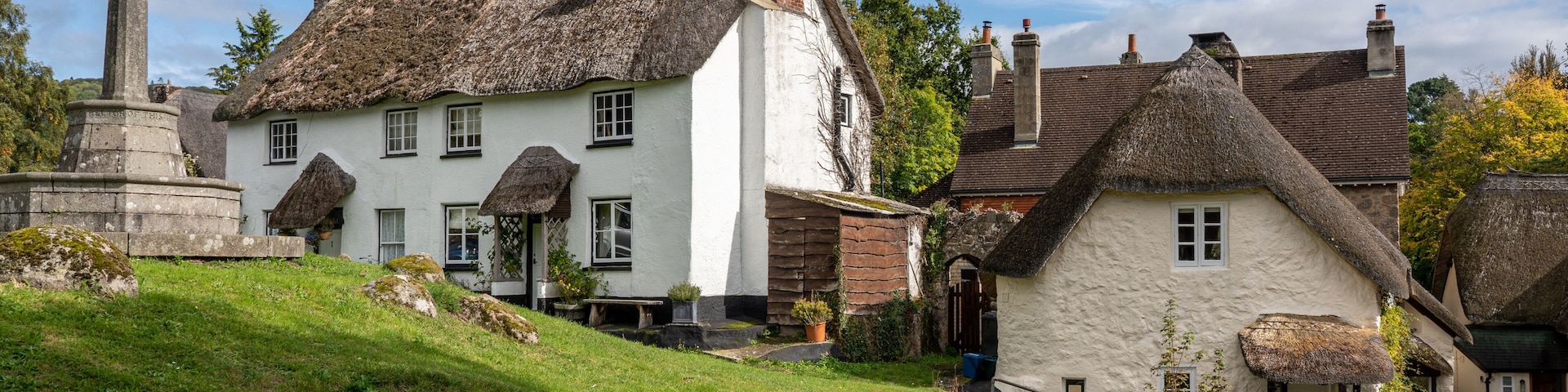 A traditional white house with a thatched roof, a stone monument, and a grassy hill.