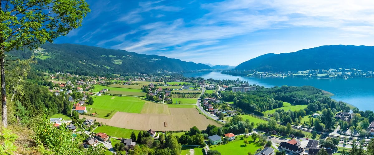 A panoramic view of a lakeside town with green fields, a river, and mountains in the background.