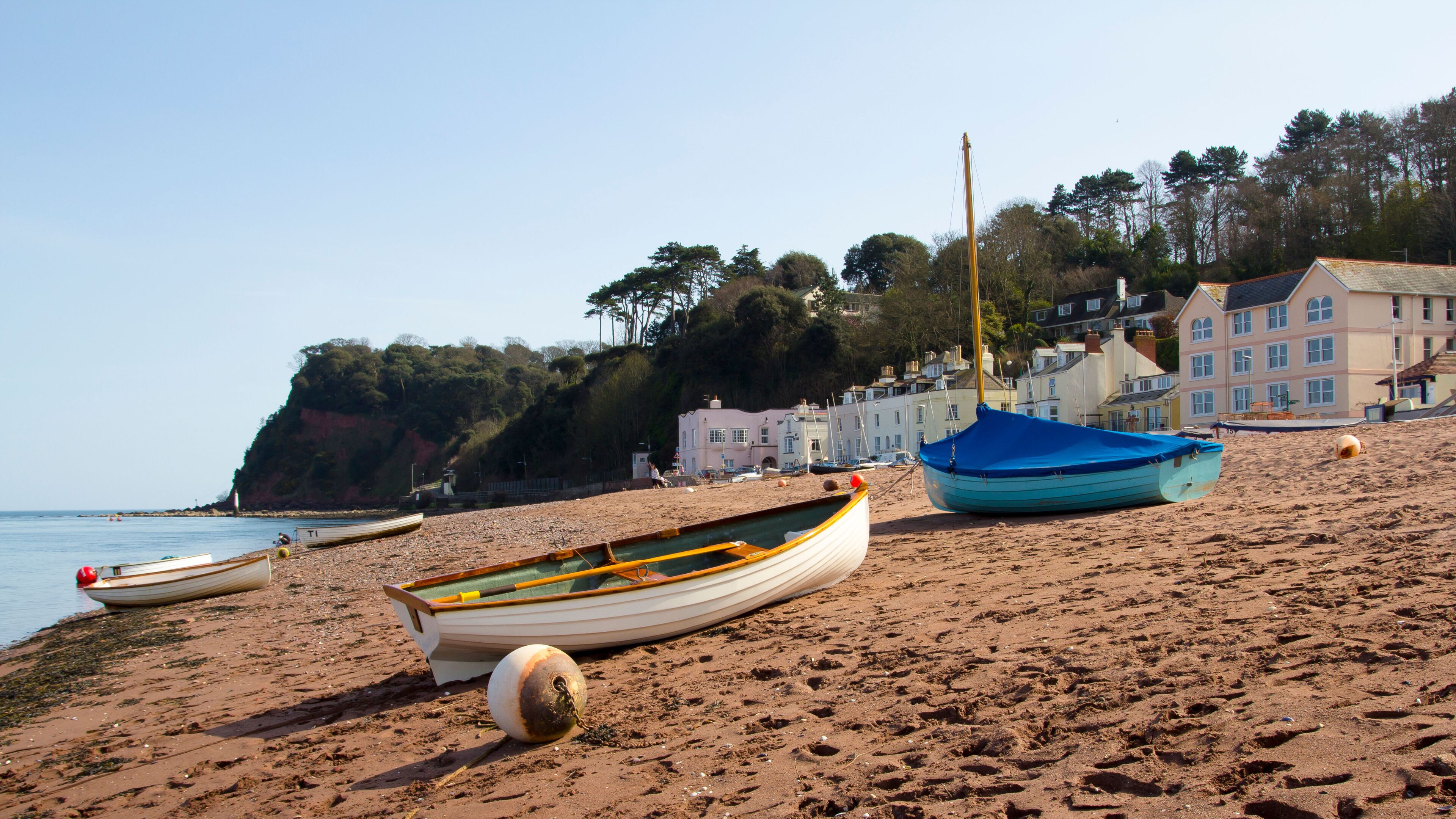 A sandy beach with several boats, a lighthouse, and houses in the background.