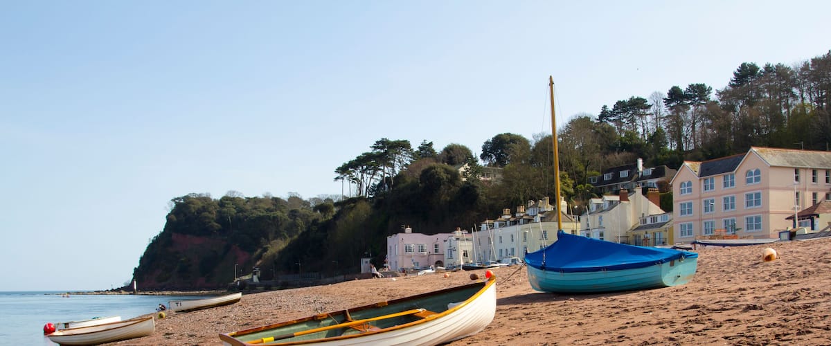 A sandy beach with several boats, a lighthouse, and houses in the background.