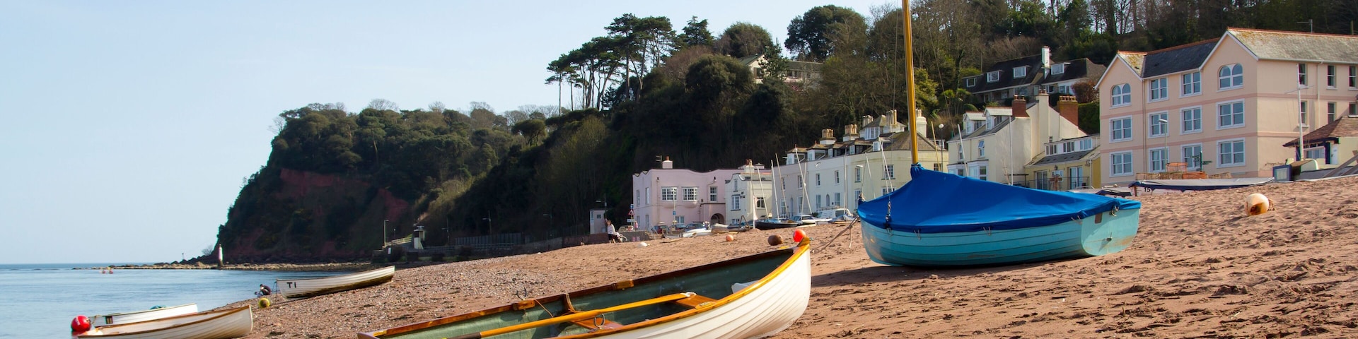 A sandy beach with several boats, a lighthouse, and houses in the background.