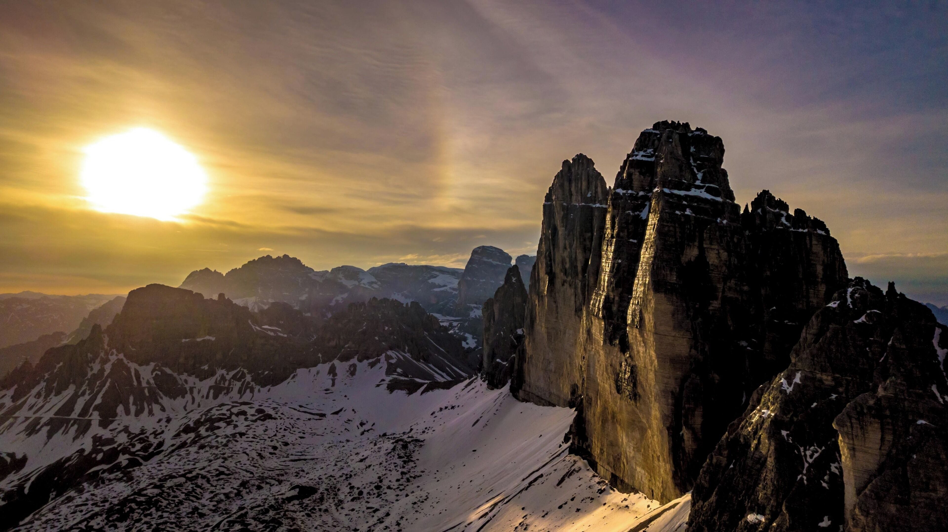 A mountain range with a prominent rock formation in the foreground and the sun setting in the background.