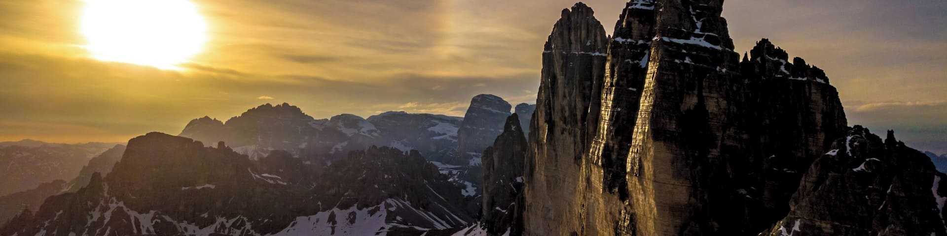 A mountain range with a prominent rock formation in the foreground and the sun setting in the background.