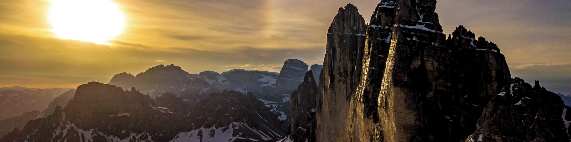 Una cadena montañosa con una formación rocosa destacada en primer plano y el sol poniéndose al fondo.