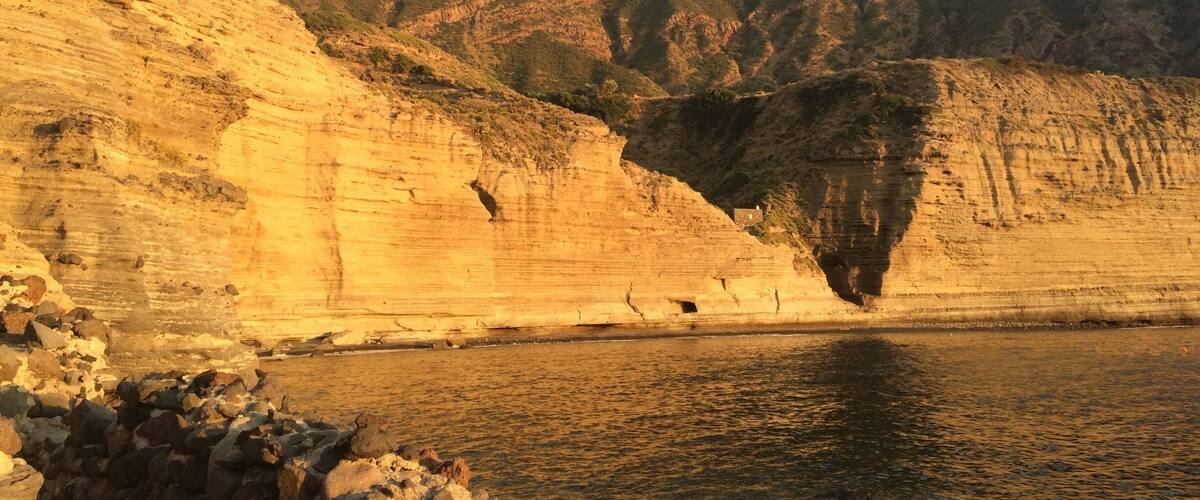A coastal cliff with a river at its base, a mountain in the background, and a partly cloudy sky.