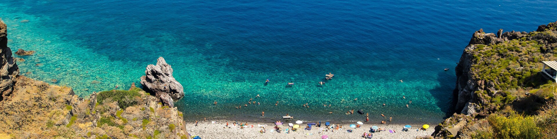 A coastal landscape with a rocky beach, clear turquoise waters, and a sailboat in the distance.