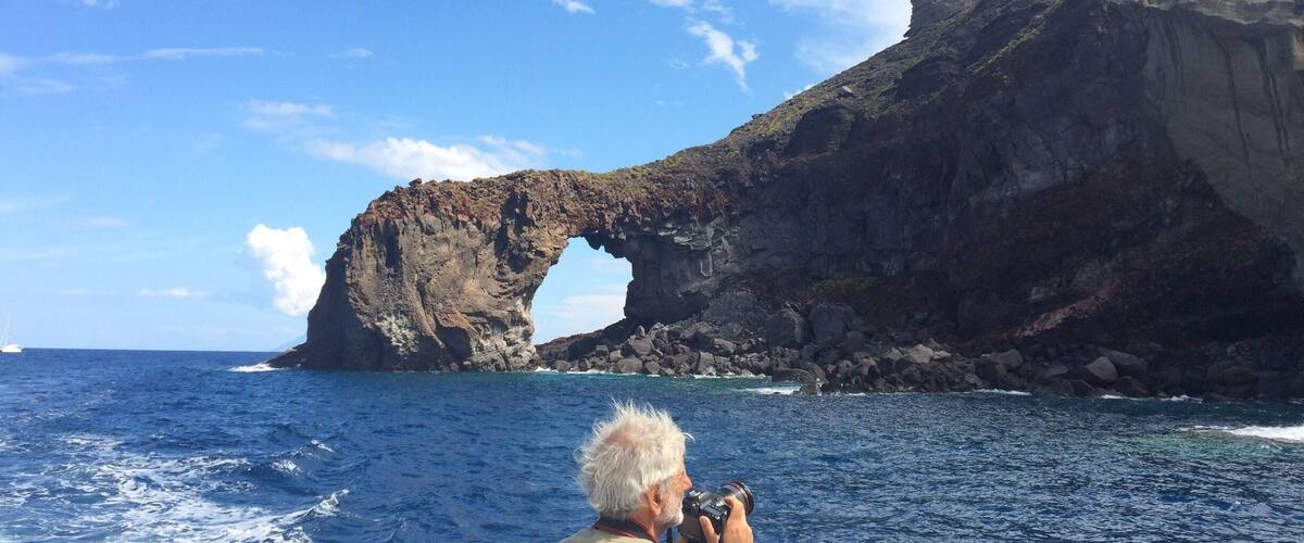 A man taking a photo of a natural rock arch formation in the ocean.