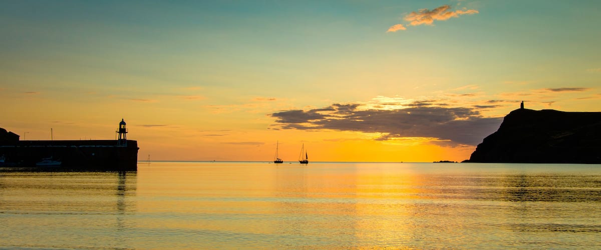 A sunset over a calm sea with a lighthouse on a cliff and a pier on the left.