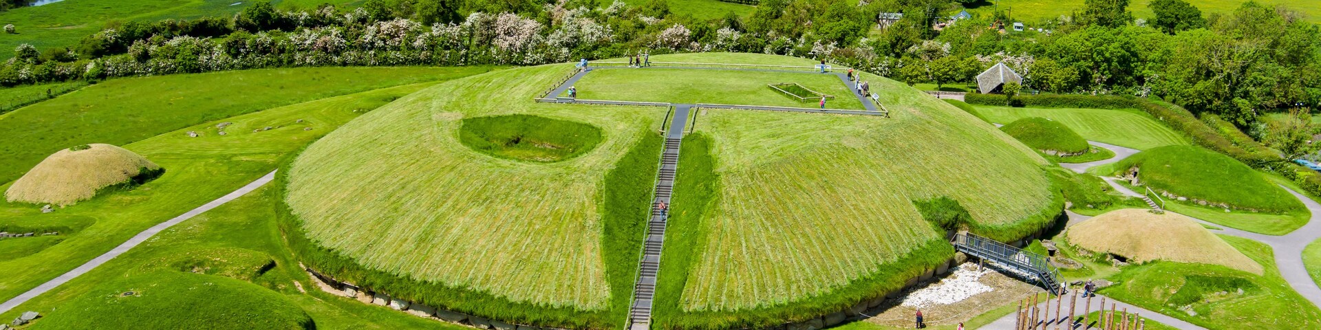Un montículo circular grande con un camino central, rodeado de campos verdes y algunos edificios dispersos.