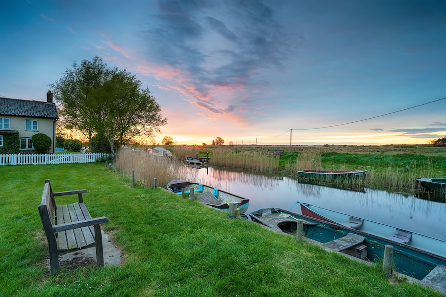 A riverside scene with a bench, boats, a house, and a sunset.