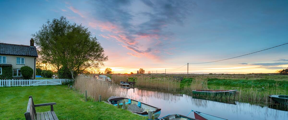 A riverside scene with a bench, boats, a house, and a sunset.
