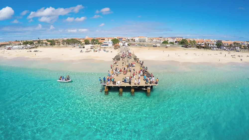 Um cais se estende sobre águas turquesa, com pessoas caminhando e uma praia ao fundo.