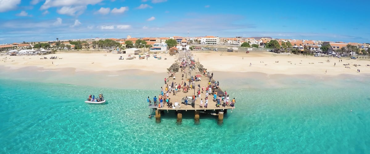 Une jetée s’avançant dans des eaux turquoise, avec des personnes qui s’y promènent, et une plage en arrière-plan.