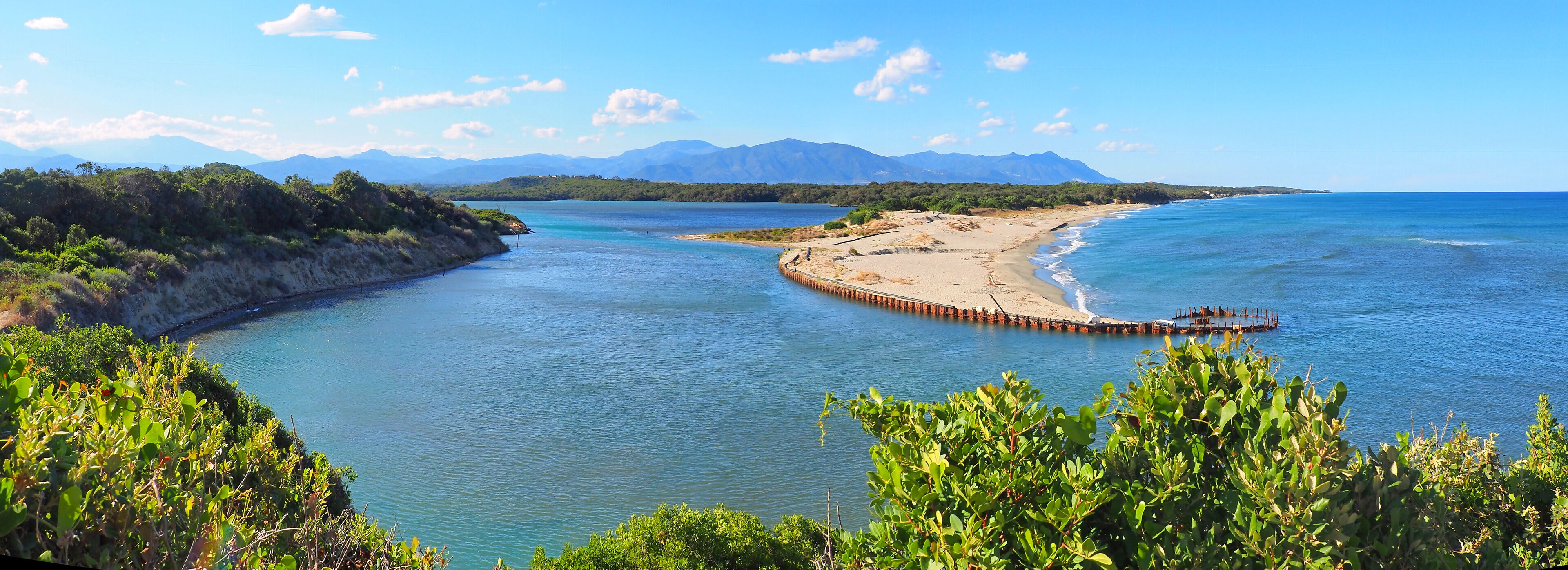 A coastal landscape with a sandy beach, a wooden pier, and mountains in the background.