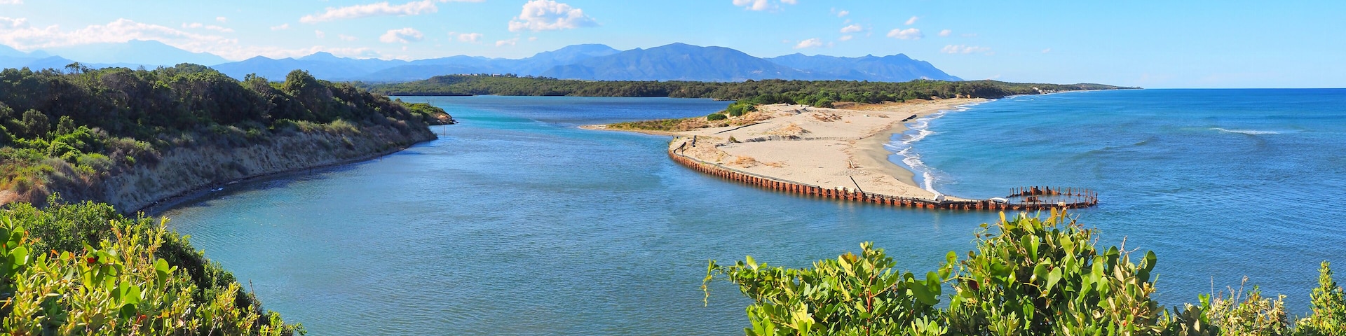 A coastal landscape with a sandy beach, a wooden pier, and mountains in the background.