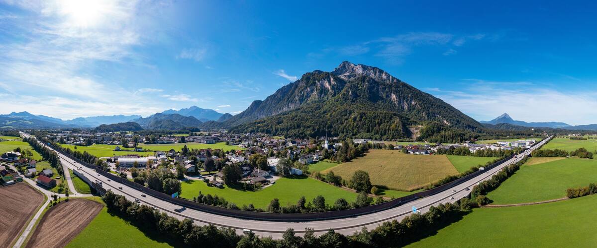 A panoramic view of a town with a prominent mountain in the background, surrounded by green fields and a highway.
