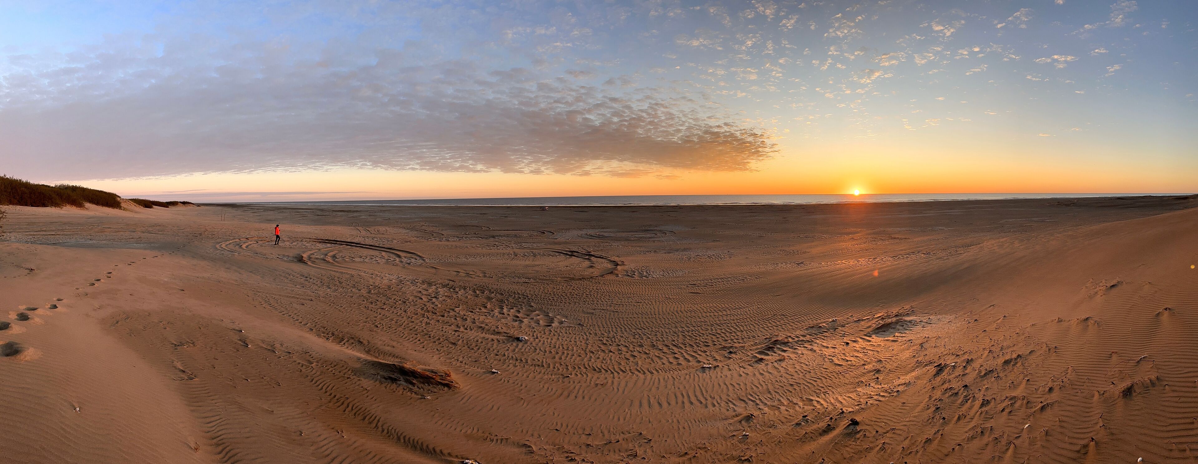 A desert landscape with a sunset over the horizon, sandy terrain with tire tracks, and sparse vegetation along the dunes.
