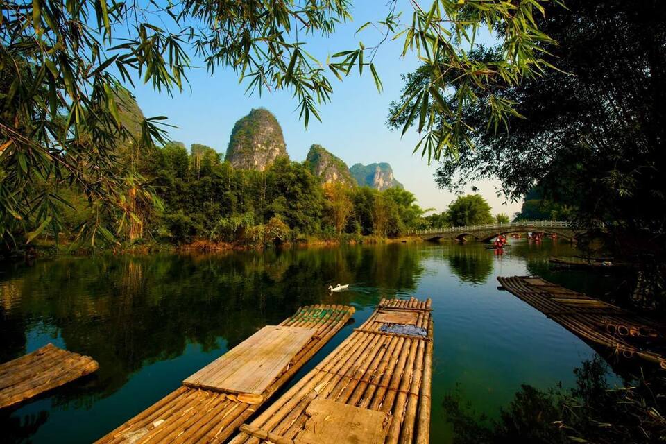A calm river with wooden rafts, lush greenery, and distant limestone karsts.