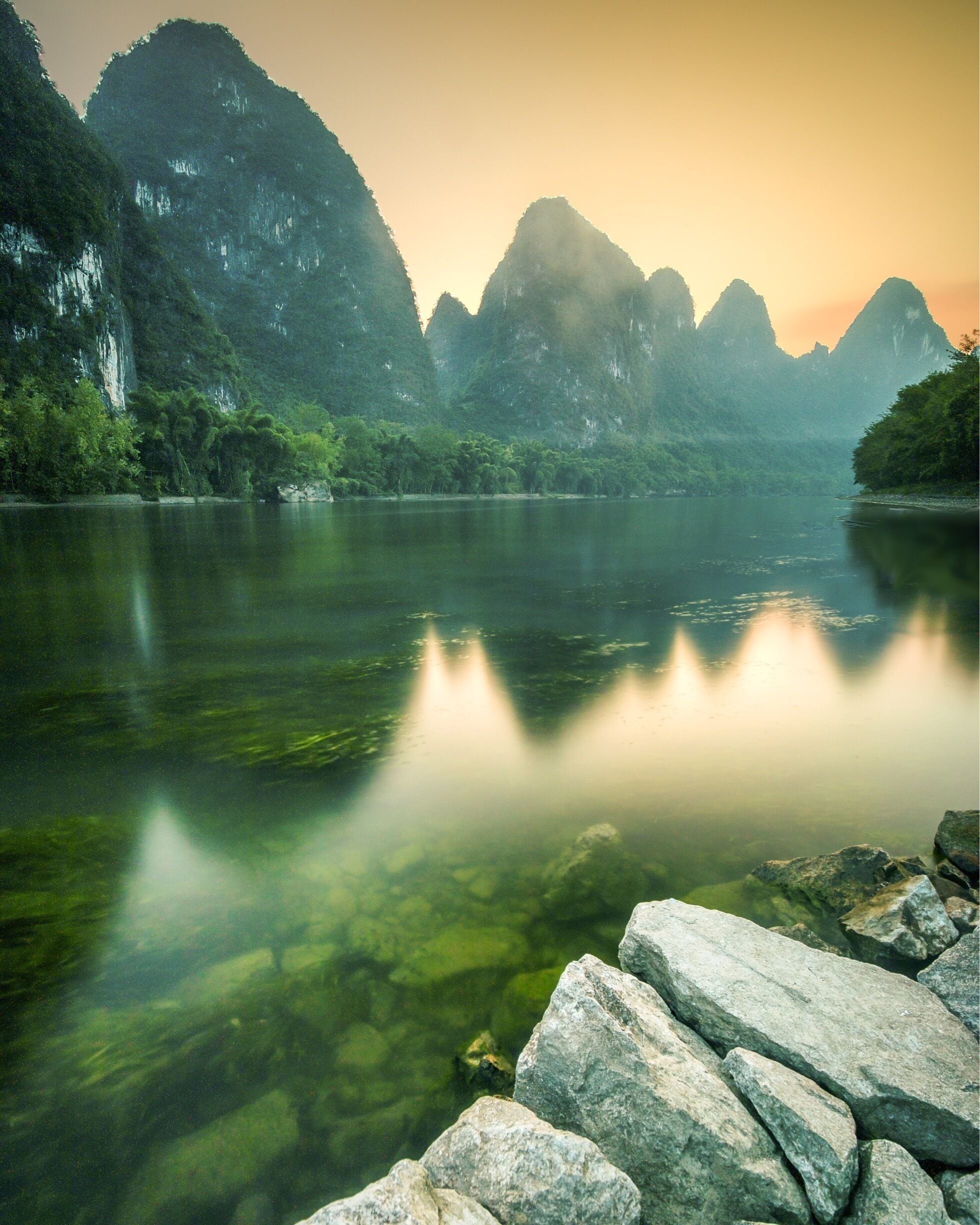A calm river with clear water reflecting the surrounding mountains and trees.