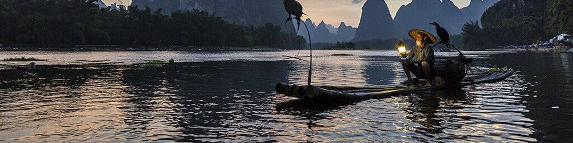 A river with a boat carrying two people and a bird, surrounded by rocky cliffs and a sunset sky.