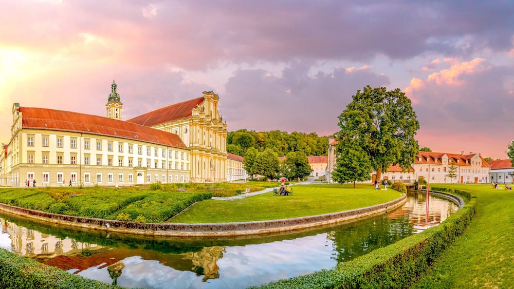 Ein historisches Gebäude mit rotem Dach, einem Kanal und einem Park mit Menschen und Bäumen.