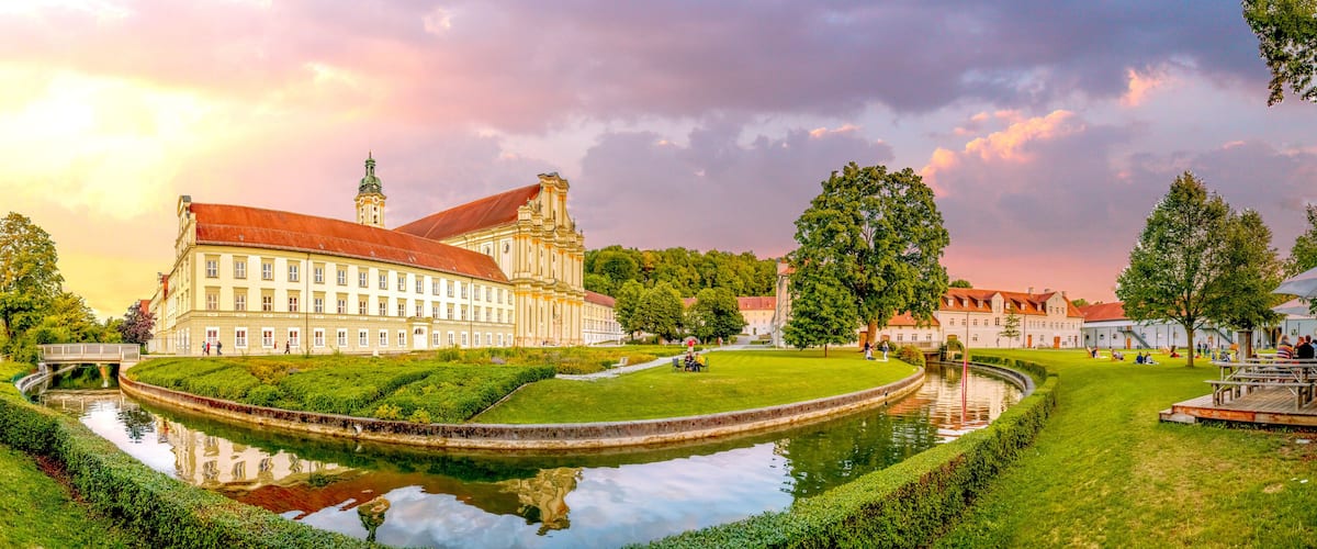 A historic building with a red roof, a canal, and a park with people and trees.