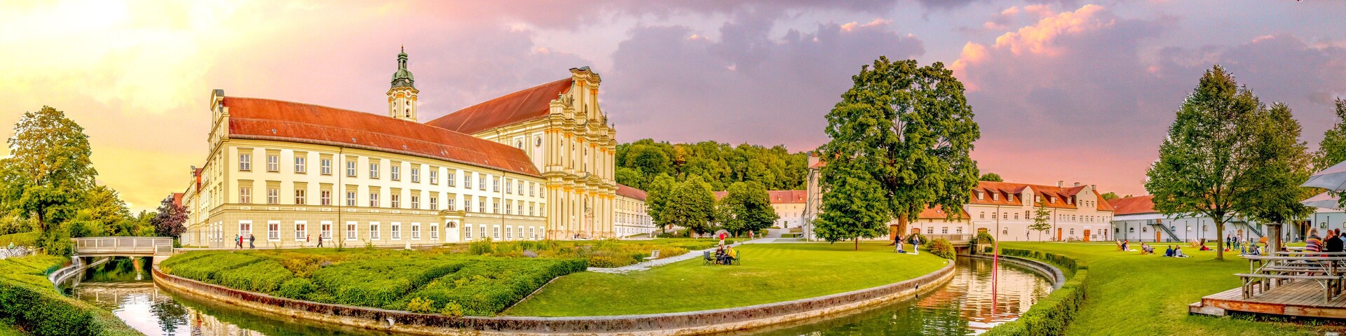 A historic building with a red roof, a canal, and a park with people and trees.