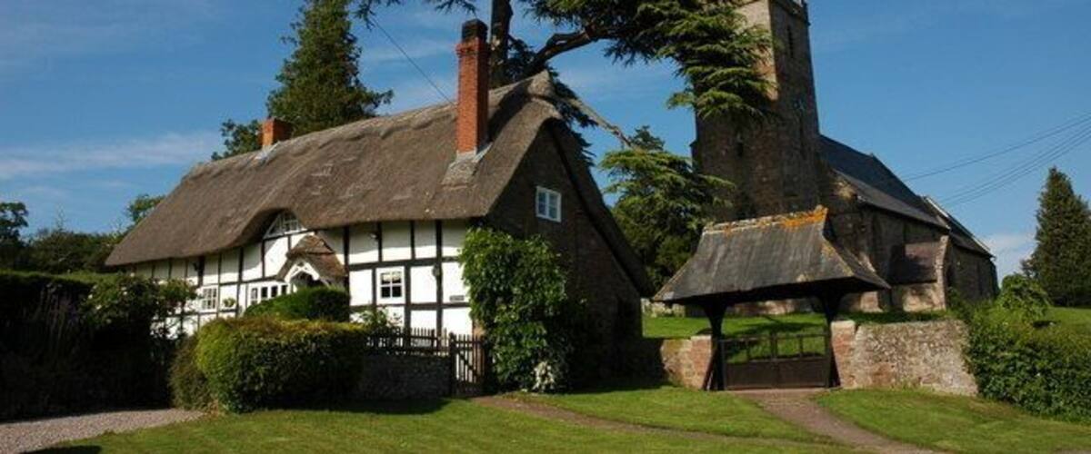 A traditional thatched-roof house with a large tree in front, a stone church with a tall steeple, and a well-maintained lawn.