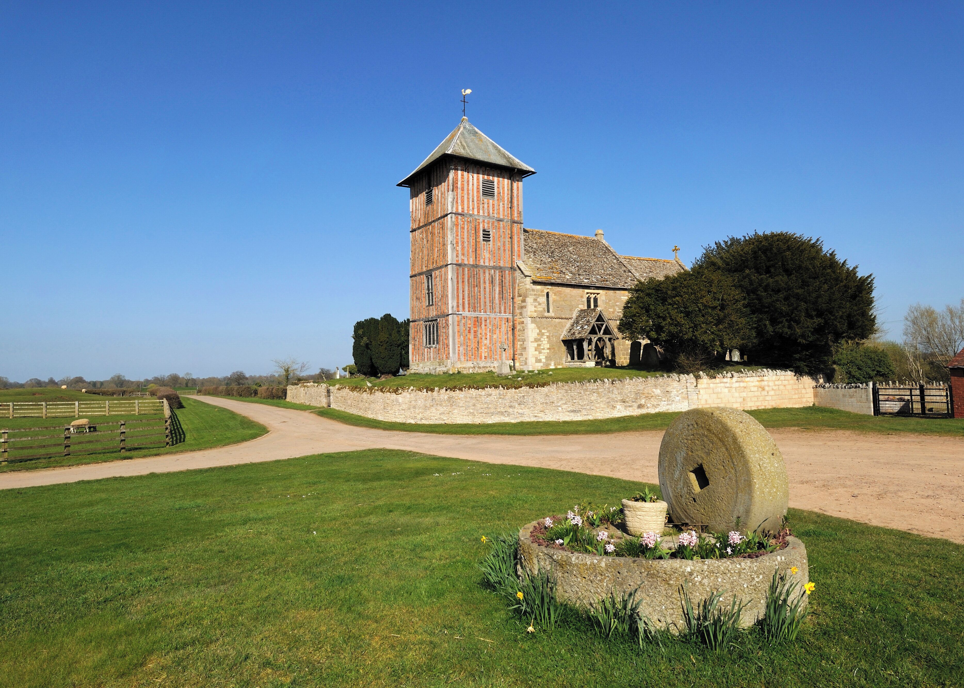 A historic brick tower with a weather vane, surrounded by a stone wall, a large tree, and a well-maintained lawn with a decorative stone feature.