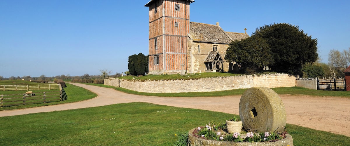A historic brick tower with a weather vane, surrounded by a stone wall, a large tree, and a well-maintained lawn with a decorative stone feature.