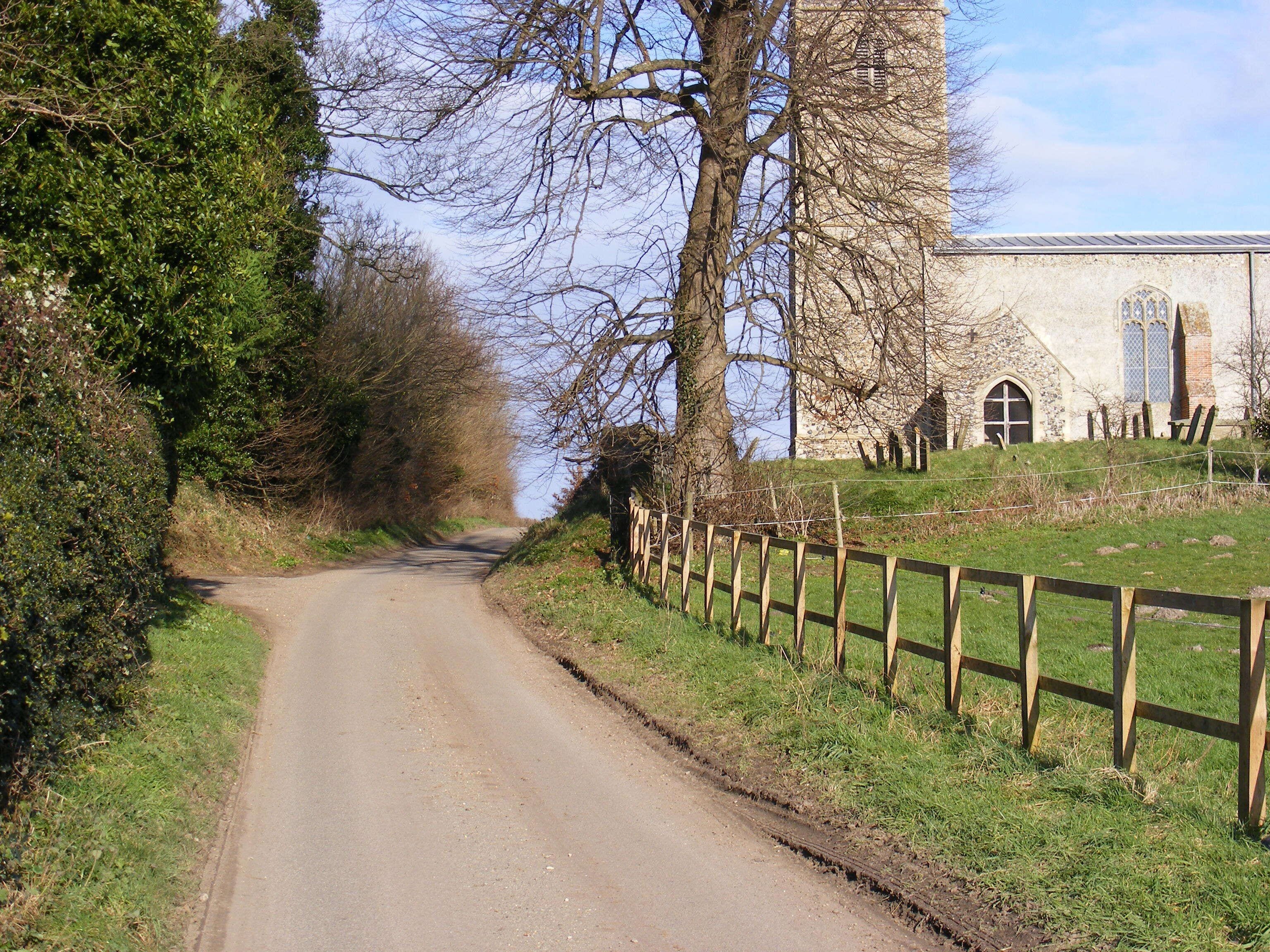 A narrow rural road flanked by trees and a stone building with a fence.