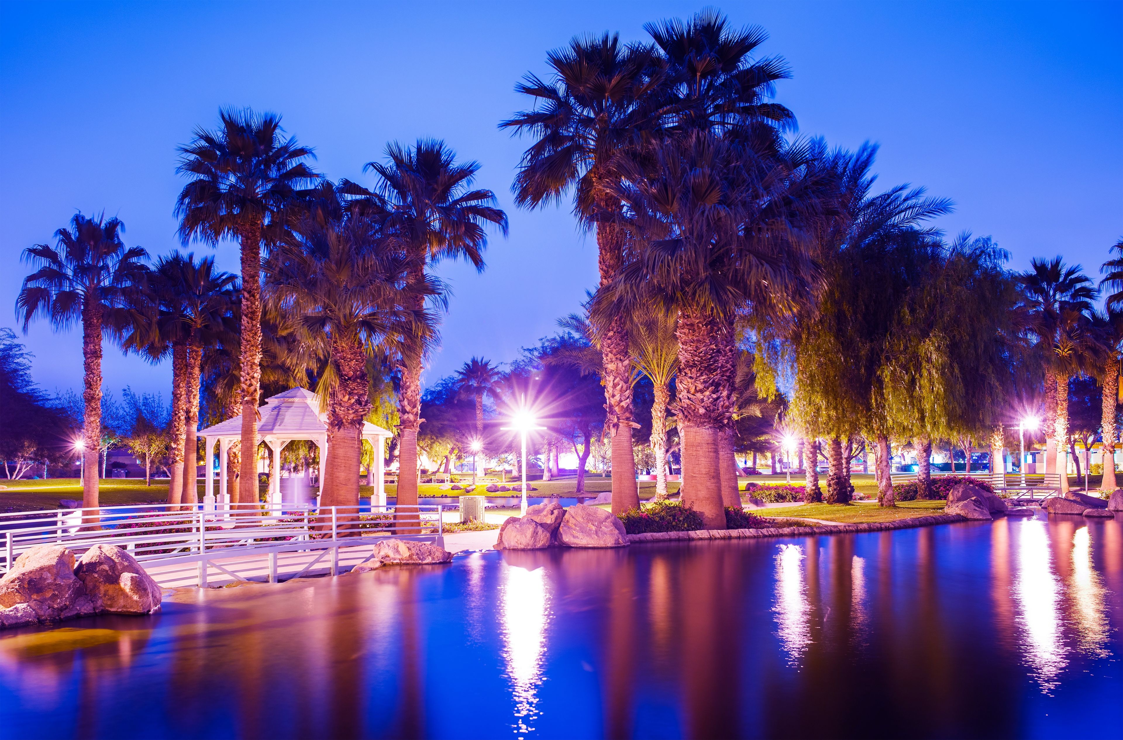 A tranquil park at dusk with palm trees, a reflecting pond, and a gazebo.