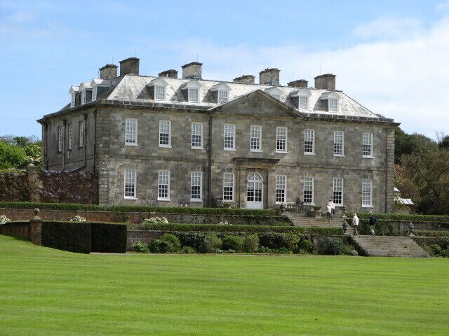 A large, historic stone building with multiple chimneys, a well-maintained lawn, and a few people walking in the foreground.