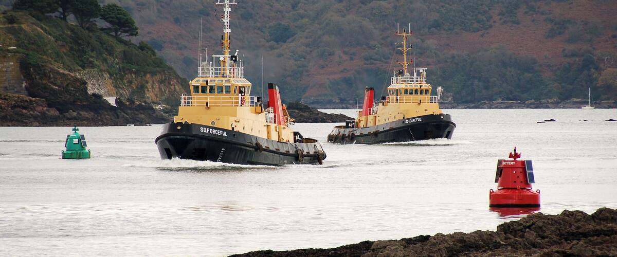 Two tugboats on a calm body of water with a rocky shoreline and hills in the background.