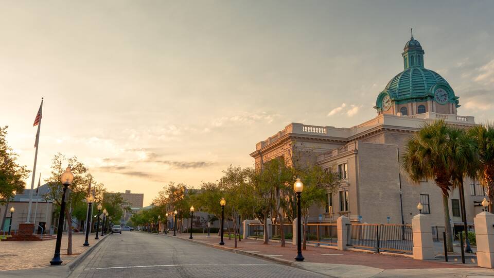 A historic building with a green dome and a clock tower, lined with palm trees and street lamps.