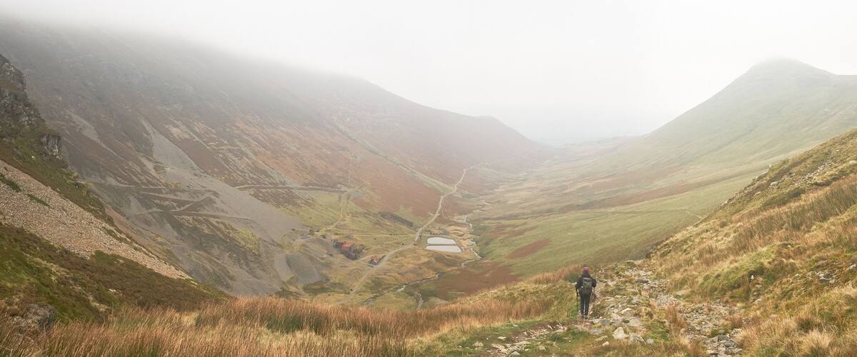 A hiker on a mountain trail with a valley and a small lake.