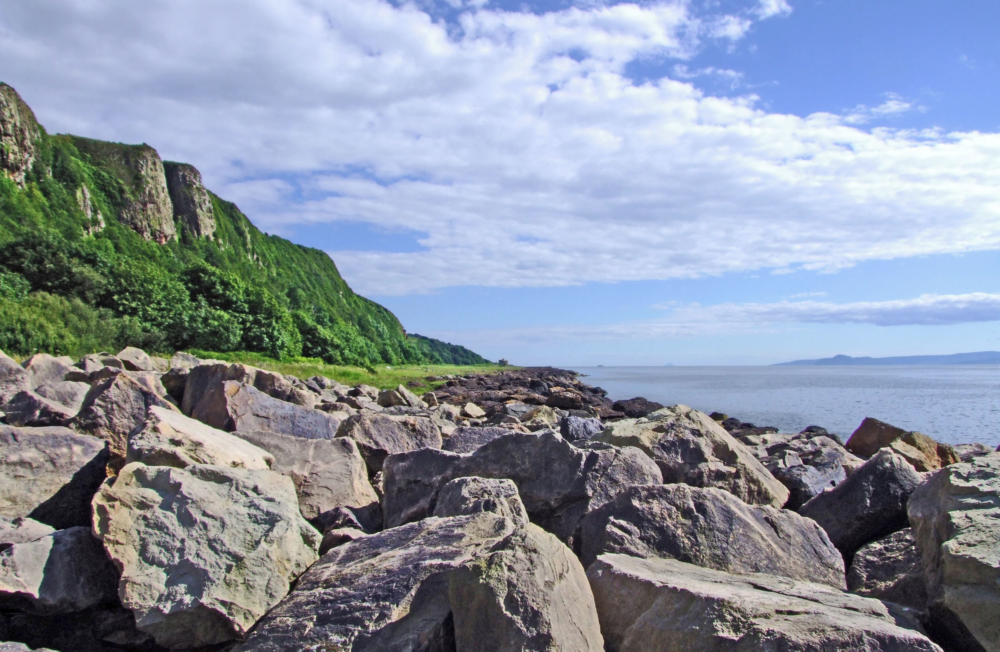 Una costa rocosa con grandes rocas y una ladera verde al fondo.