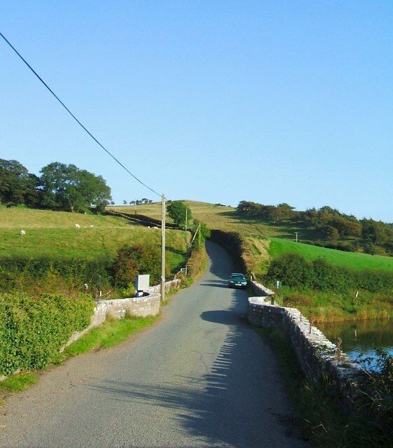 A narrow rural road flanked by fields and a stone wall.