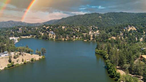 aerial shot of a gorgeous summer landscape with rippling lake water, green trees, grass and plants with homes on the hillside, blue sky, cloud sand a rainbow at Lake Gregory in Crestline California