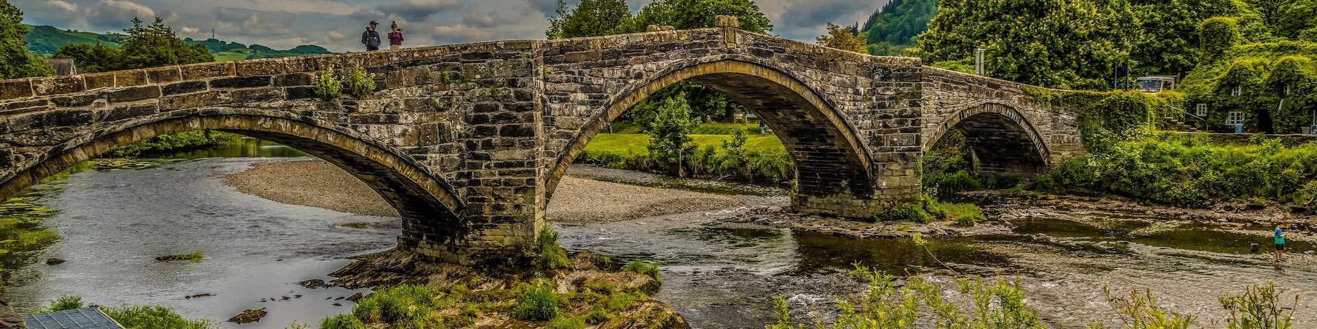 A stone bridge over a river with greenery and hills in the background.