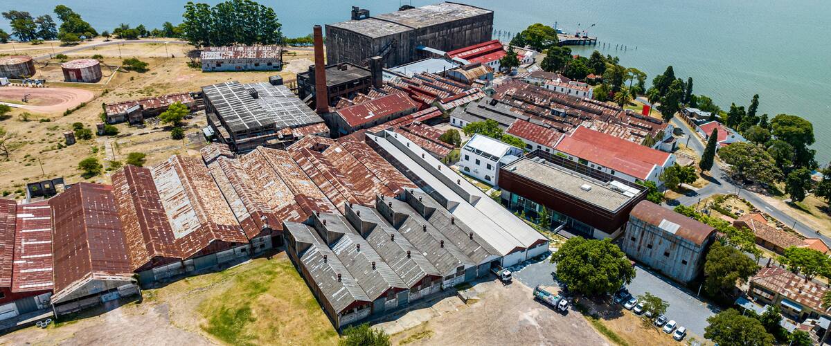 An industrial area with warehouses, a large building with a rusted roof, and a body of water in the background.