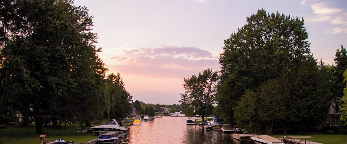 A calm river with boats docked on both sides, surrounded by trees and a sunset sky.