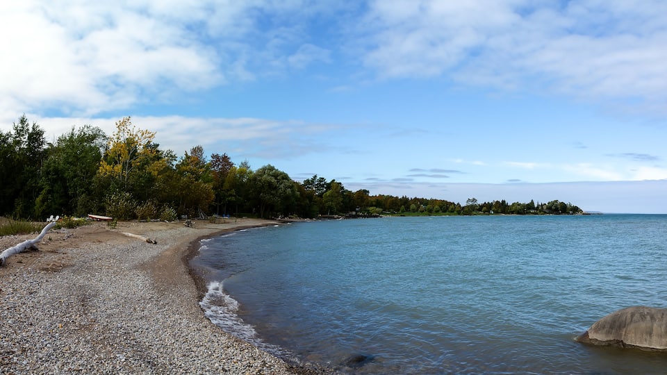 A pebble beach with a large rock in the water, surrounded by trees and a house.