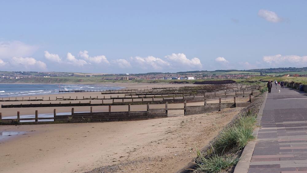 A coastal walkway with a wooden fence, beach, and distant buildings.
