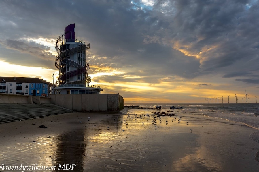 Escena costera con una turbina eólica, un muelle y una puesta de sol.