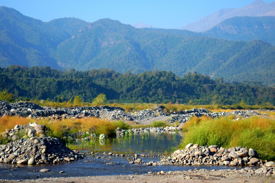 Un río con orillas rocosas y una cadena montañosa boscosa al fondo.