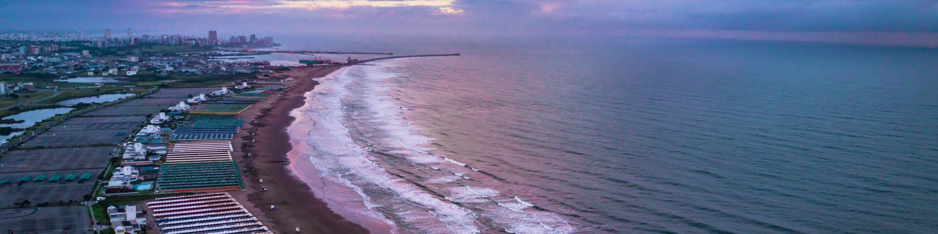 Vista aérea de uma praia com fileiras de tendas e um horizonte urbano ao fundo.